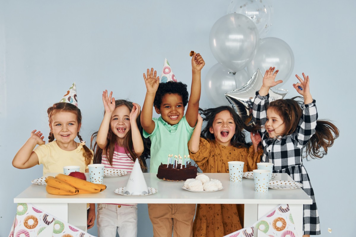 Sits by the table. Children on celebrating birthday party indoors have fun together.